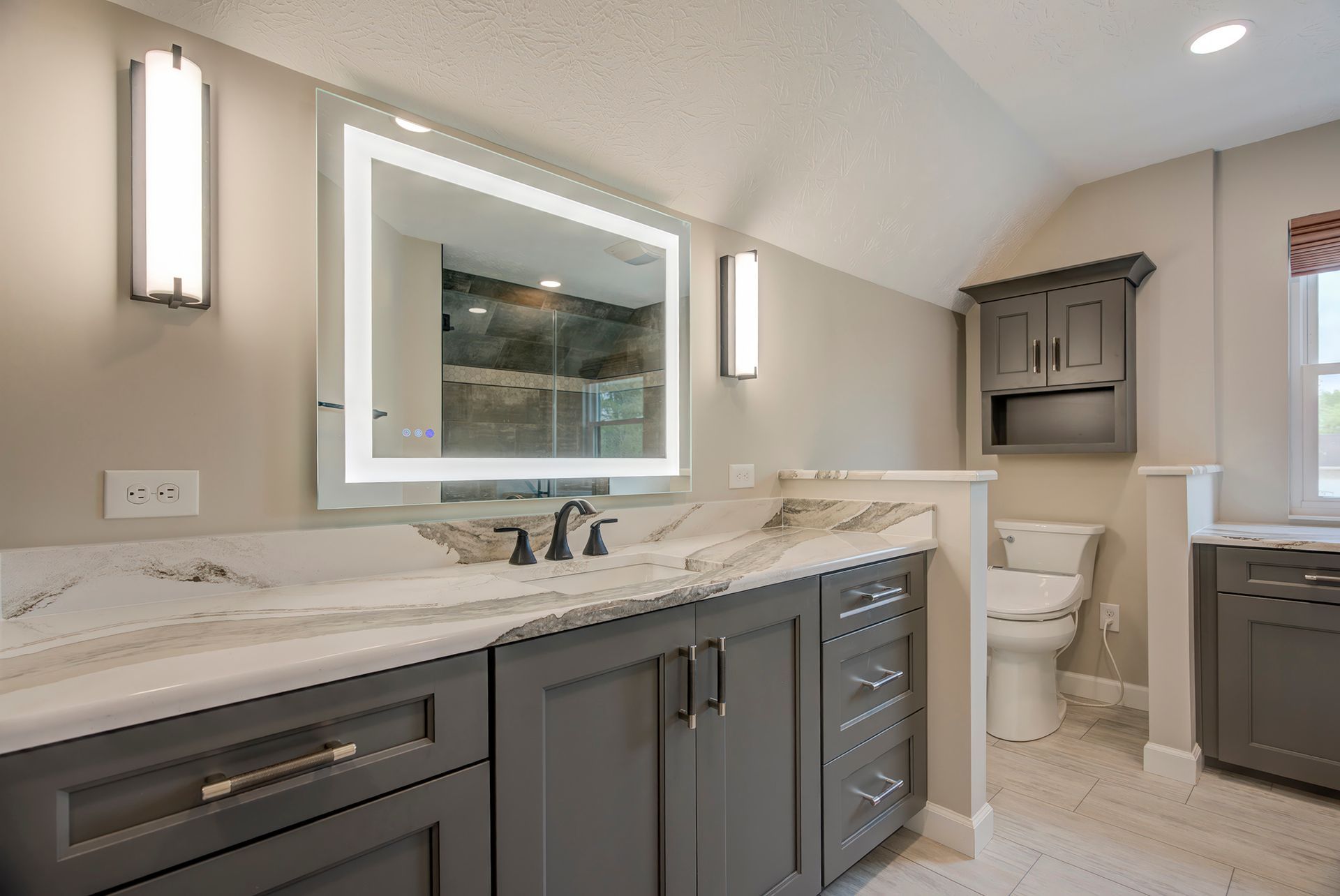Modern gray bathroom with vanity, lit mirror, toilet, and storage cabinet.