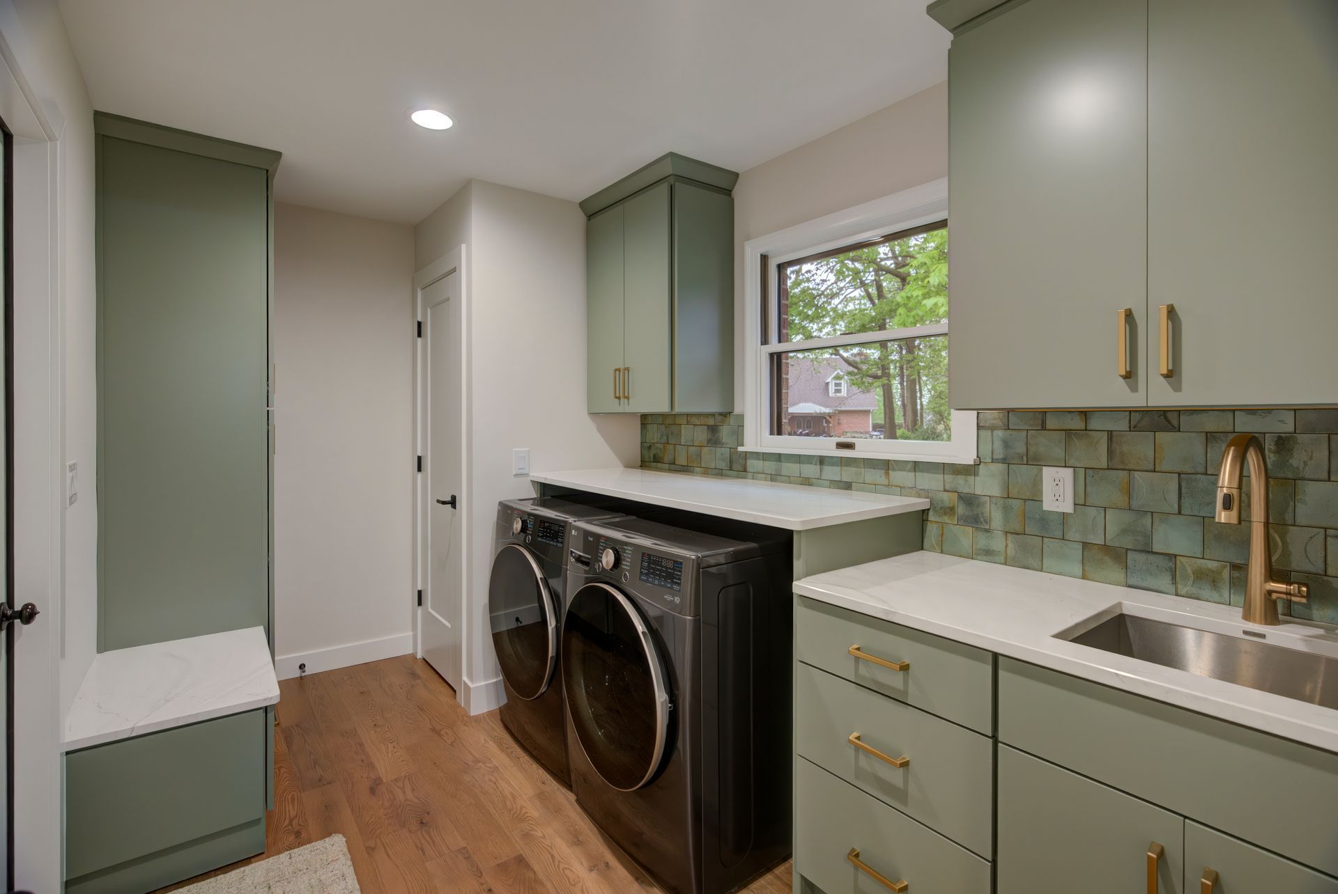 Green and white laundry room with washer, dryer, sink, cabinets, window, and hardwood floors.