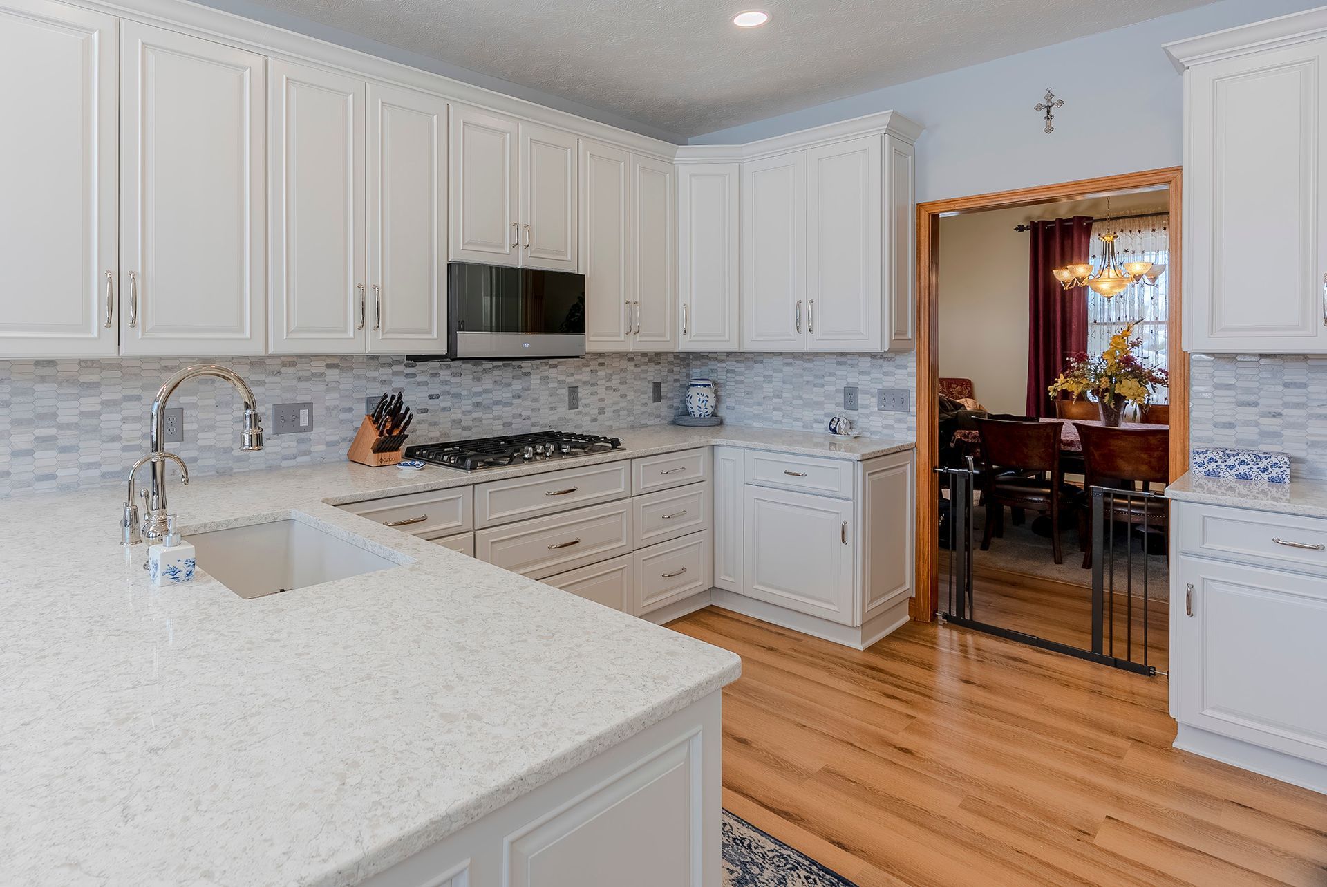 White kitchen with light countertops, cabinetry, backsplash, and hardwood floors. Dining room visible in the background.
