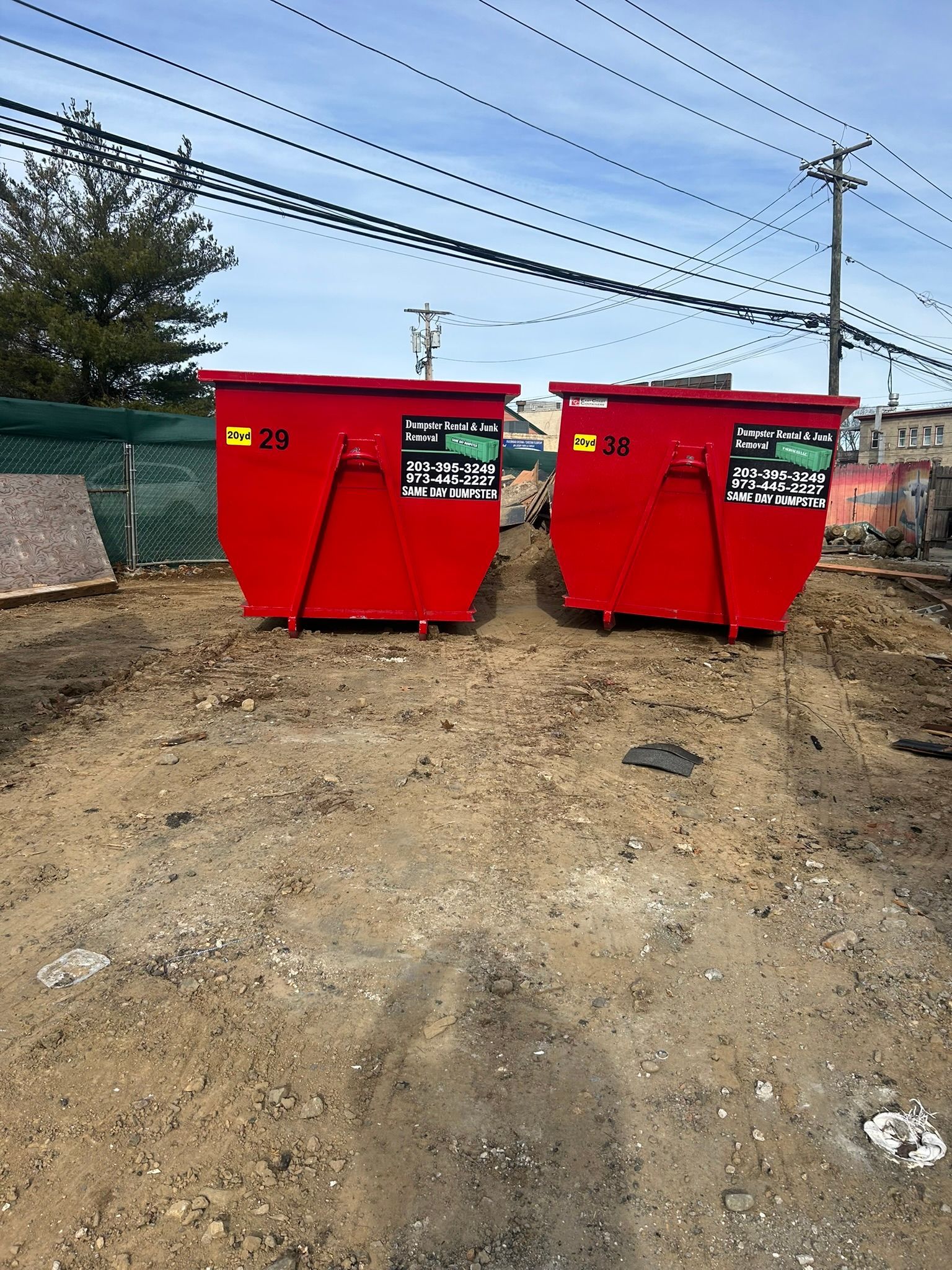 Two red dumpsters side-by-side on a dirt lot, beneath power lines.