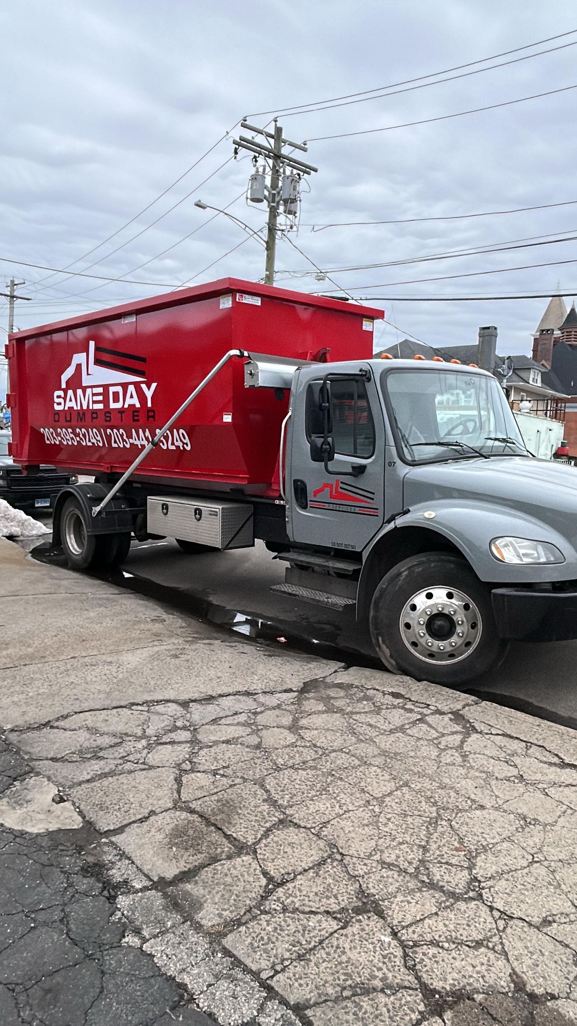 Red dumpster on a gray truck parked on a paved street. The truck has the logo "Same Day" on the side.