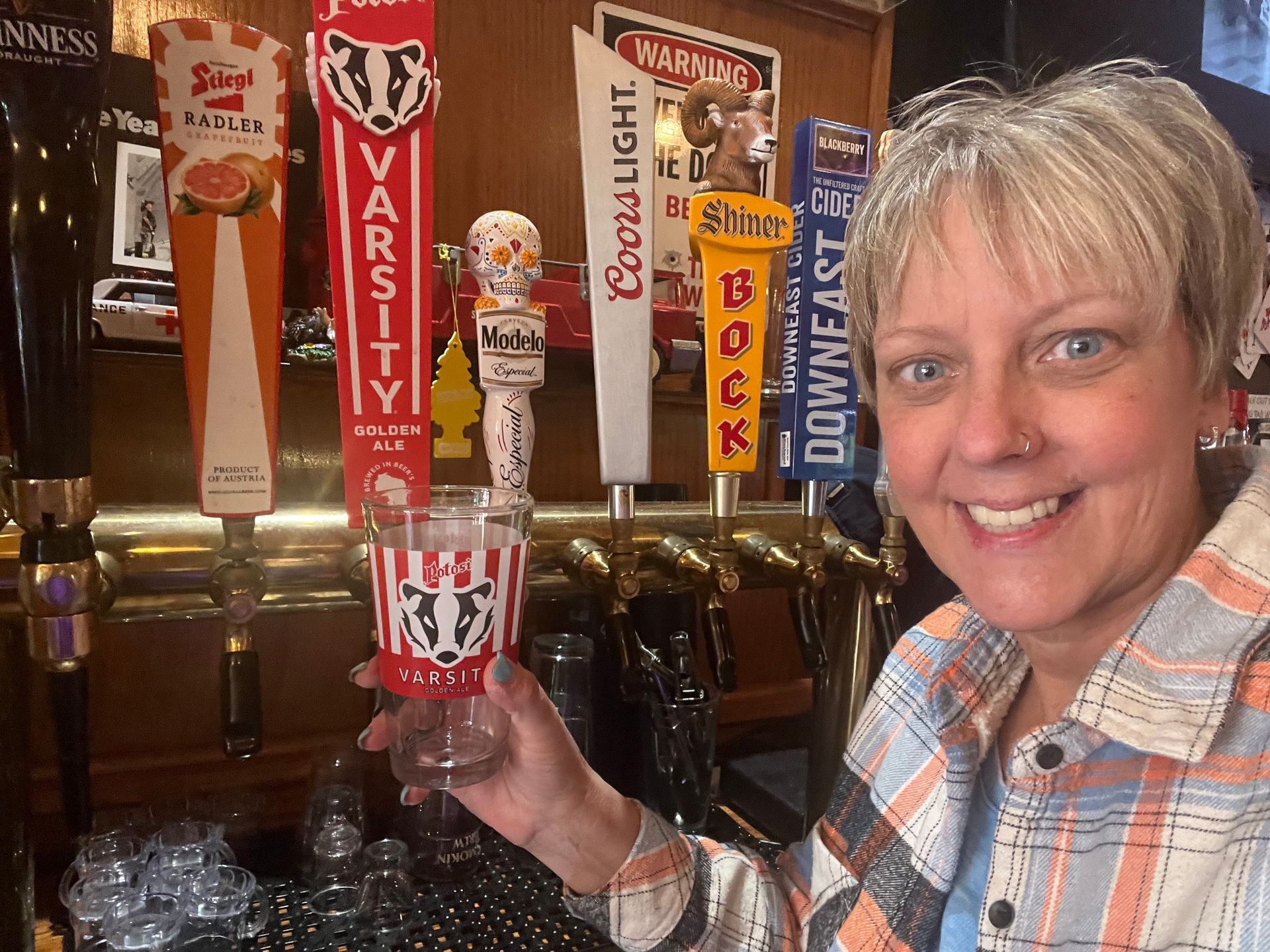 A woman is holding a glass of beer in front of a beer tap that says varsity