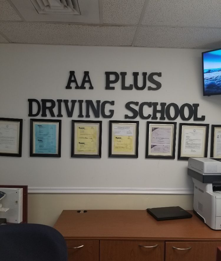 AA Plus Driving School sign on a white wall above a desk with framed documents.