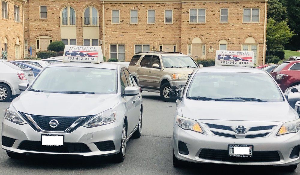 Two silver sedans parked in front of a brick building. Both cars have signs on their roofs with a phone number. Other parked cars and a beige SUV are visible.