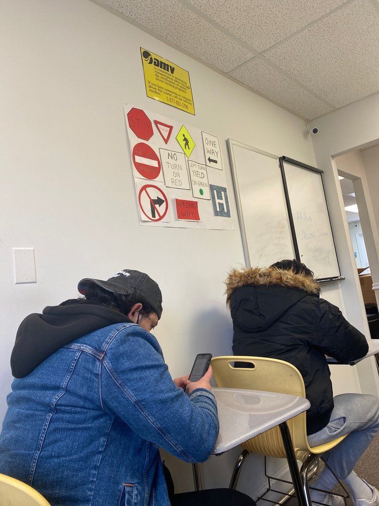 Two students sitting at desks in a classroom, looking at their phones. Road signs and a whiteboard are mounted on the wall.
