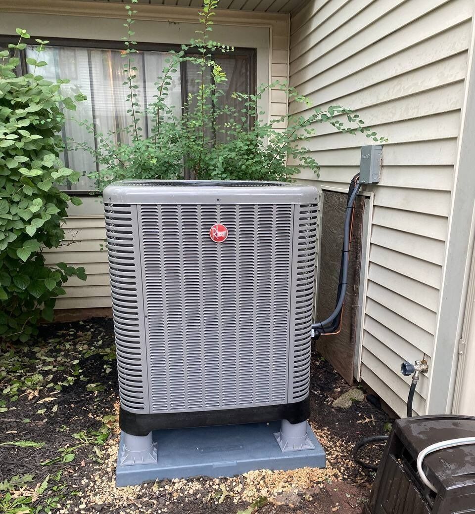 Gray Rheem AC unit with bushes on top, beside a house with white siding.