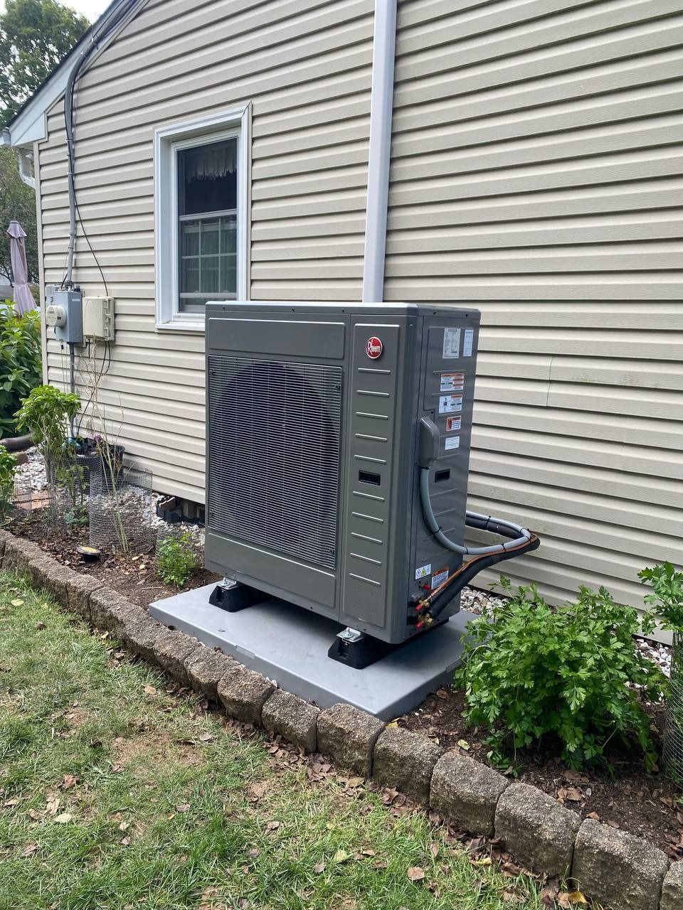 Rheem heat pump unit on a concrete pad next to a house with beige siding and a small window.