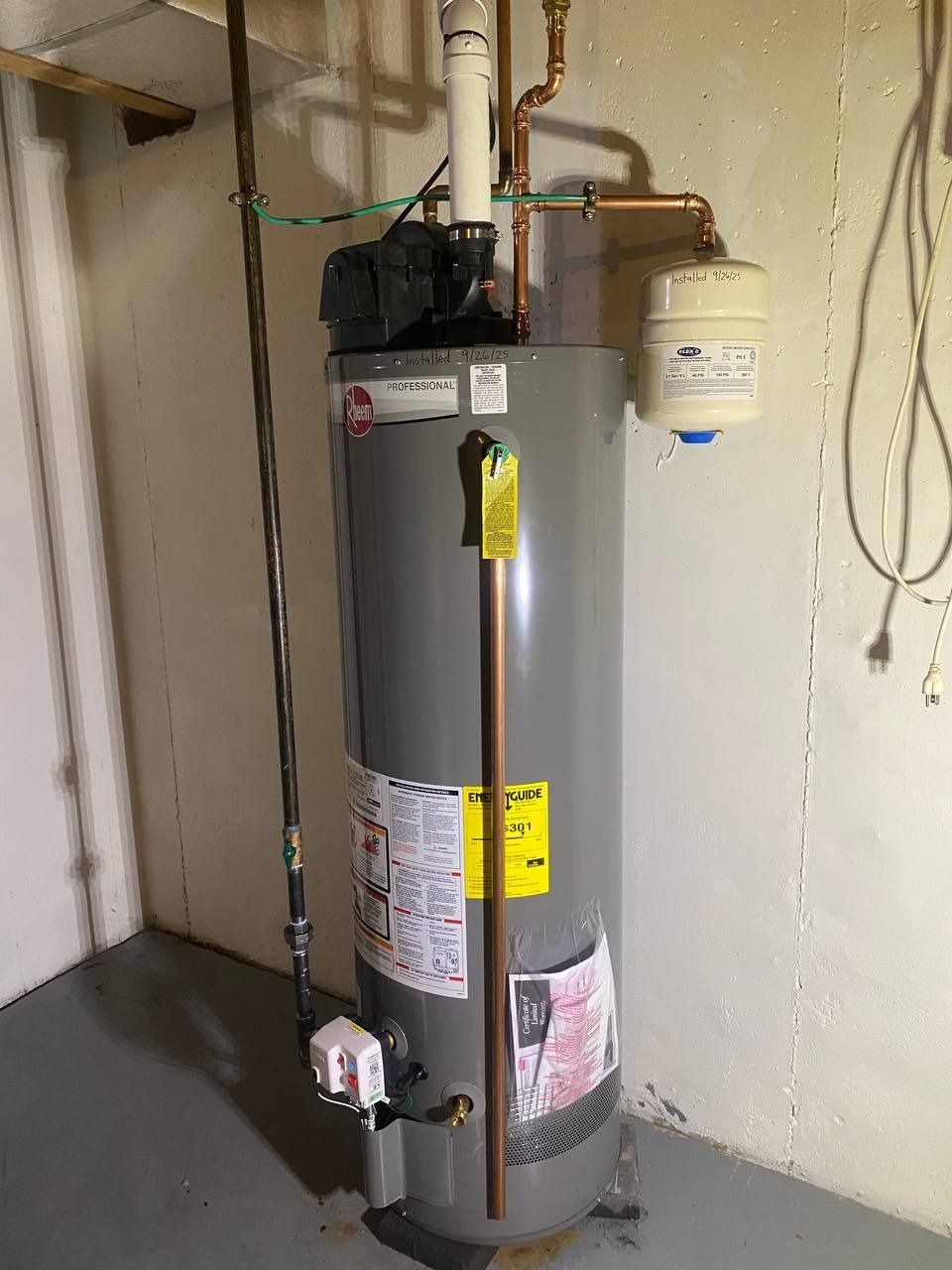 Water heater in a basement corner, with pipes and a pressure tank attached.