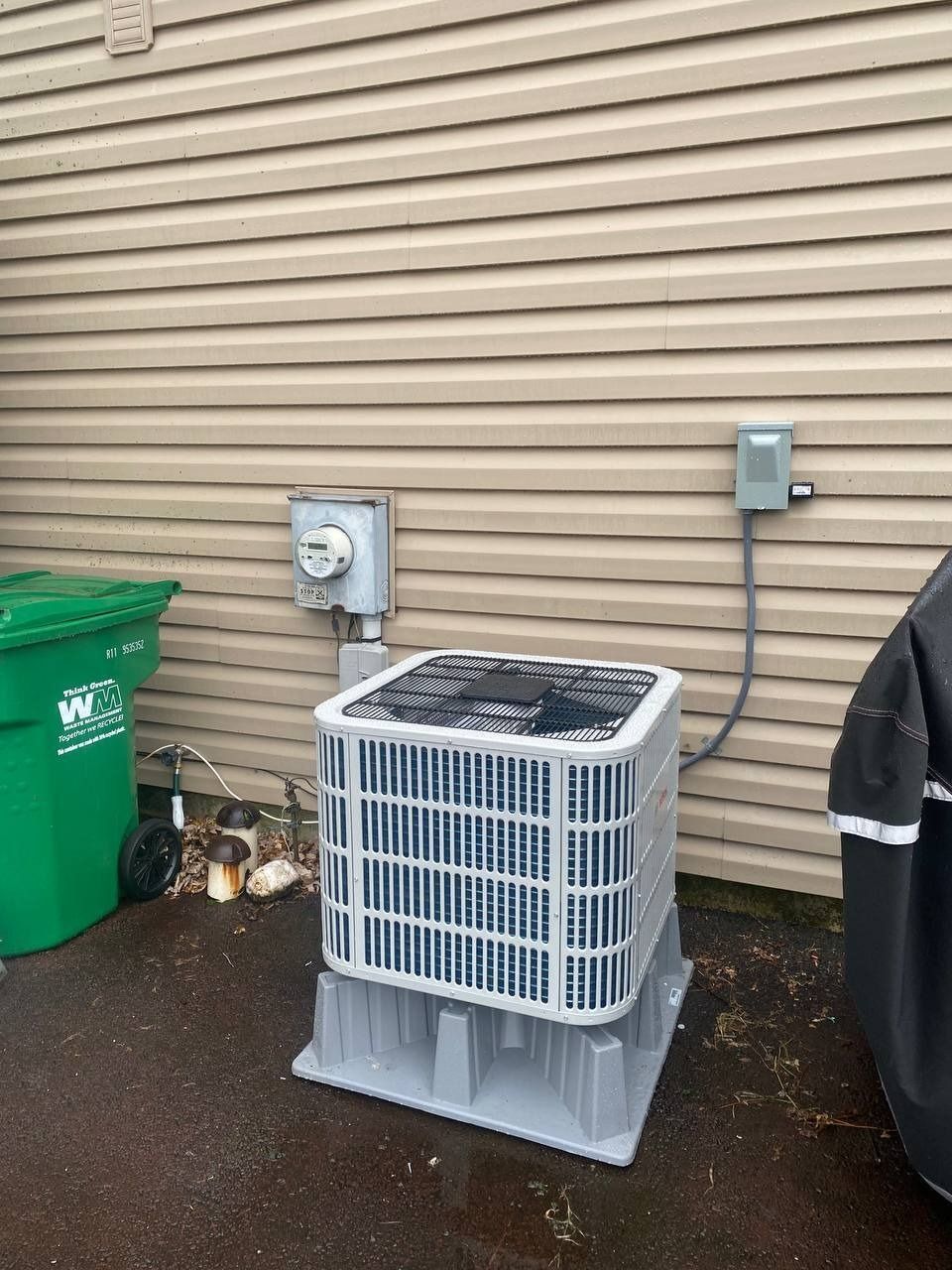 Air conditioner on a gray stand next to a beige house, green trash can, and electrical box.