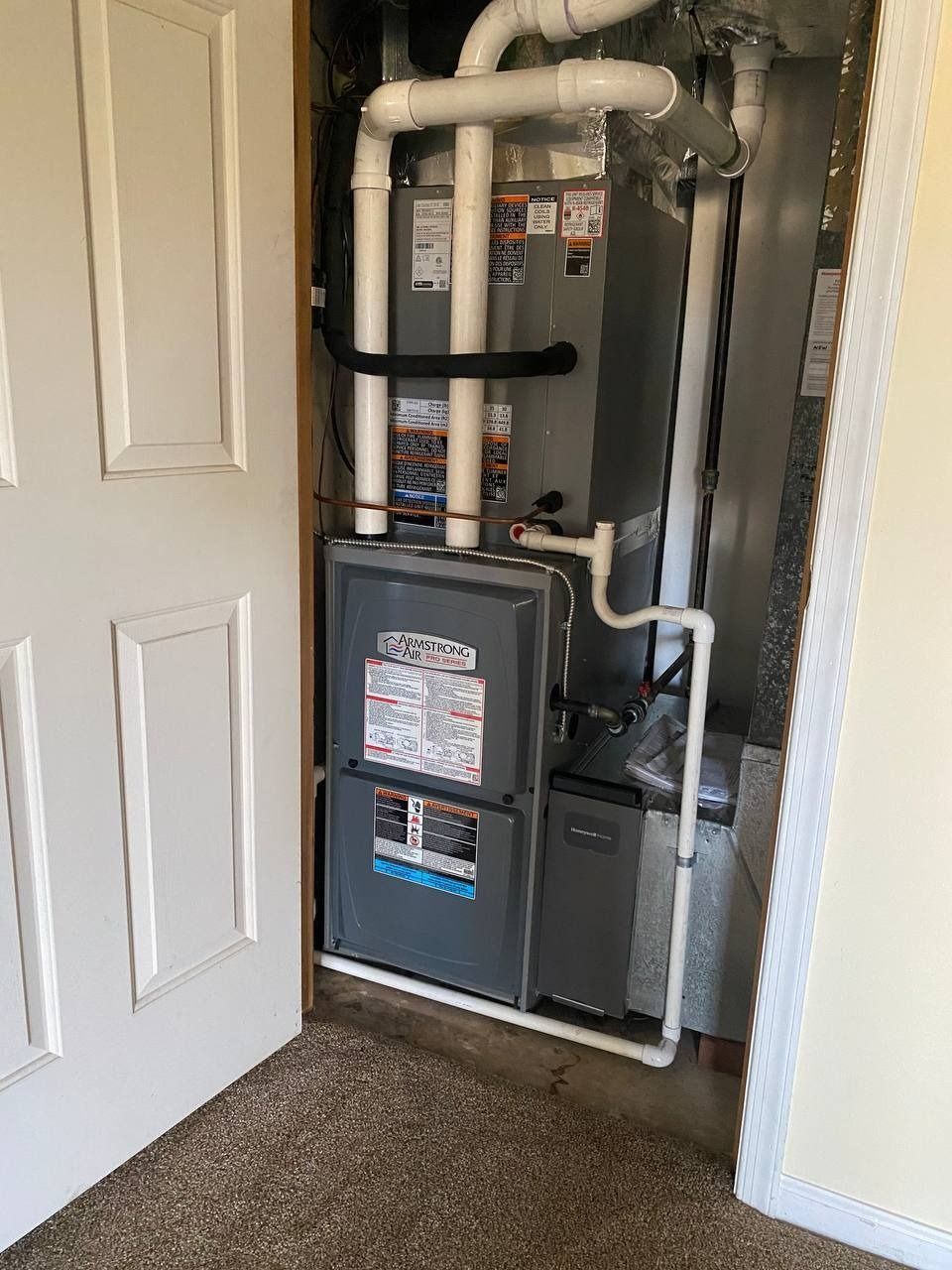 Furnace in a closet, with white pipes, gray metal unit, and a partially open white door.