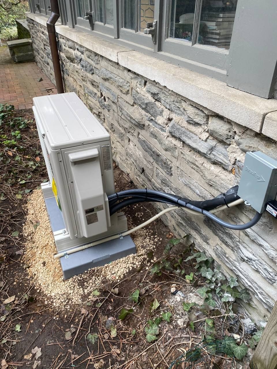 An outdoor HVAC unit on a gravel base next to a stone building wall with windows.