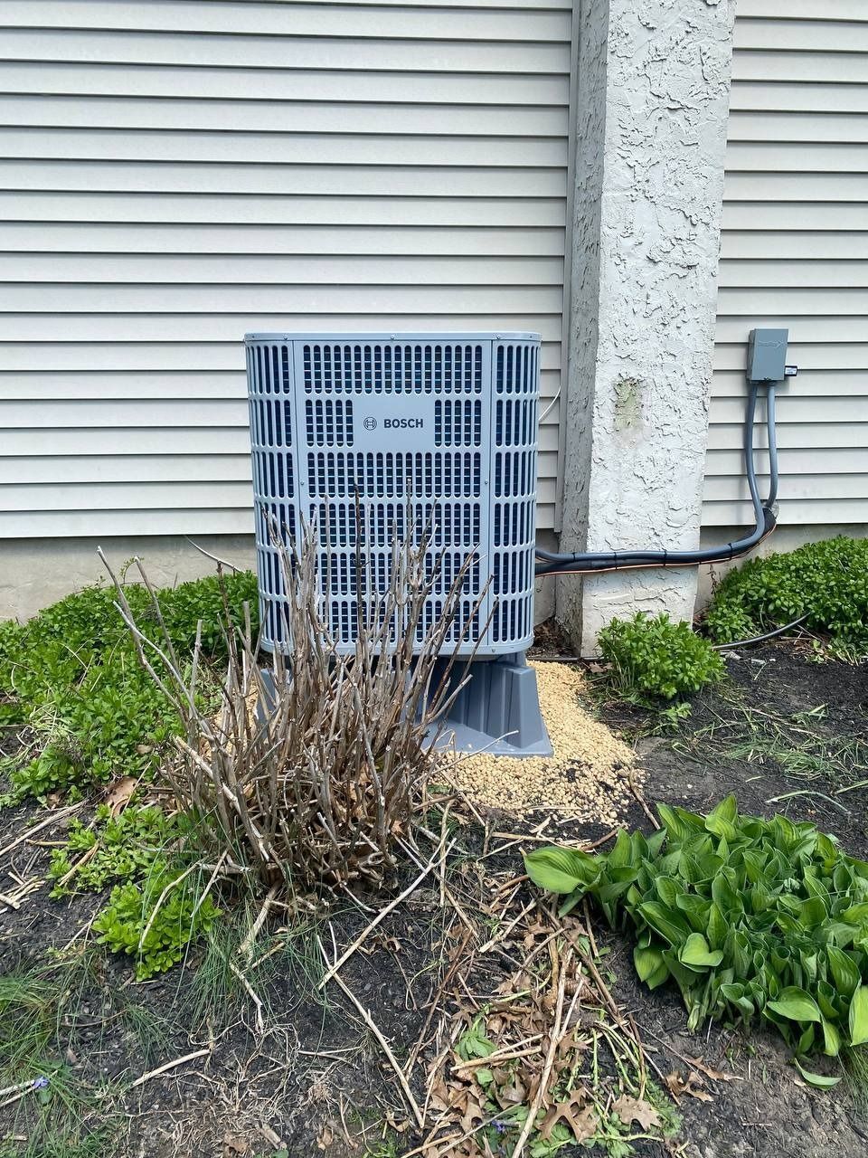 Gray air conditioning unit outside a building with light siding, surrounded by landscaping.