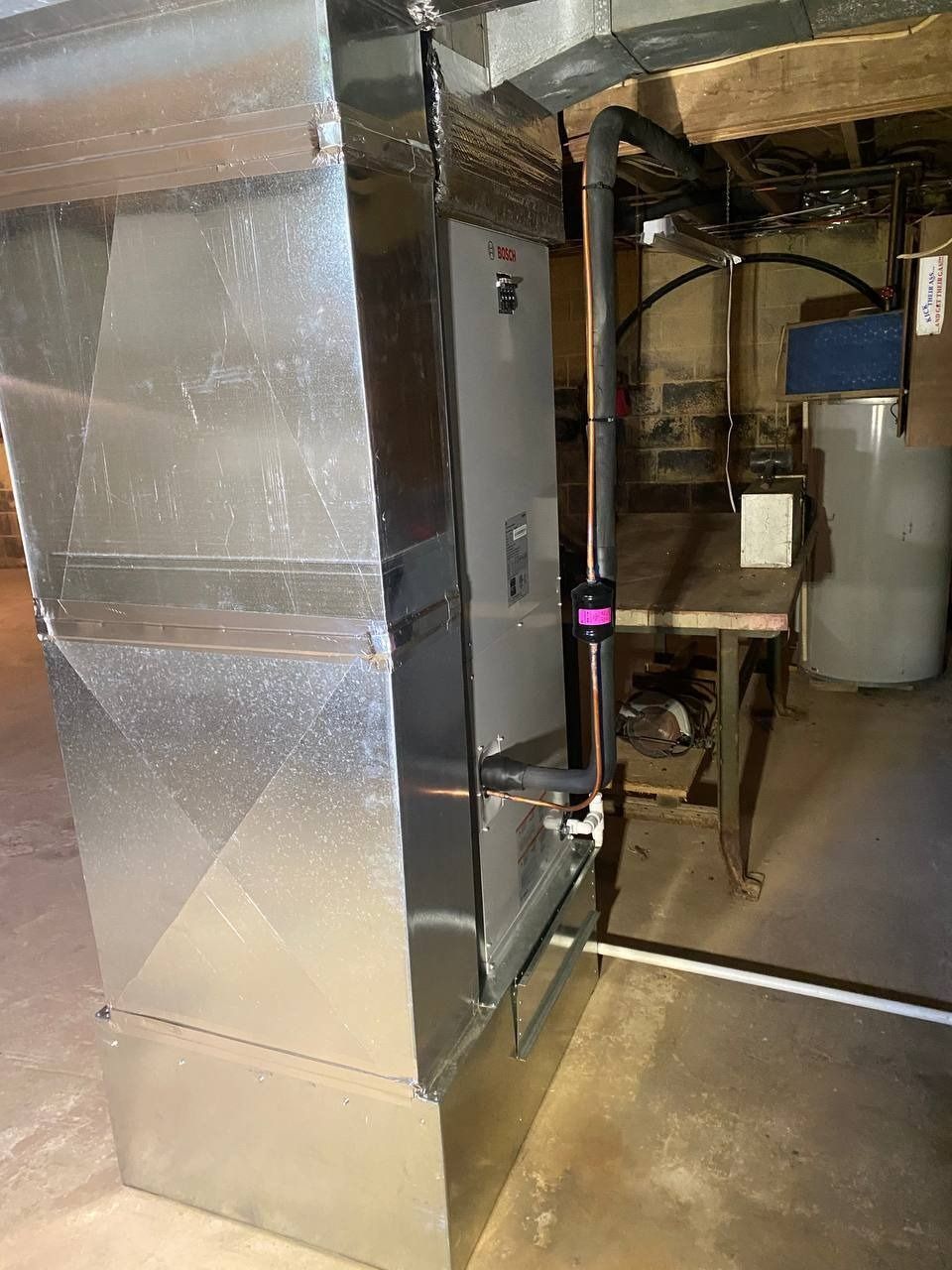 A furnace in a basement with silver ductwork, black pipe and a white tank.