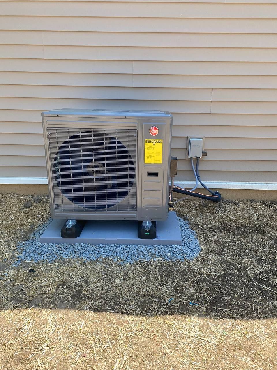 Gray Rheem heat pump on a gravel base next to a beige house.
