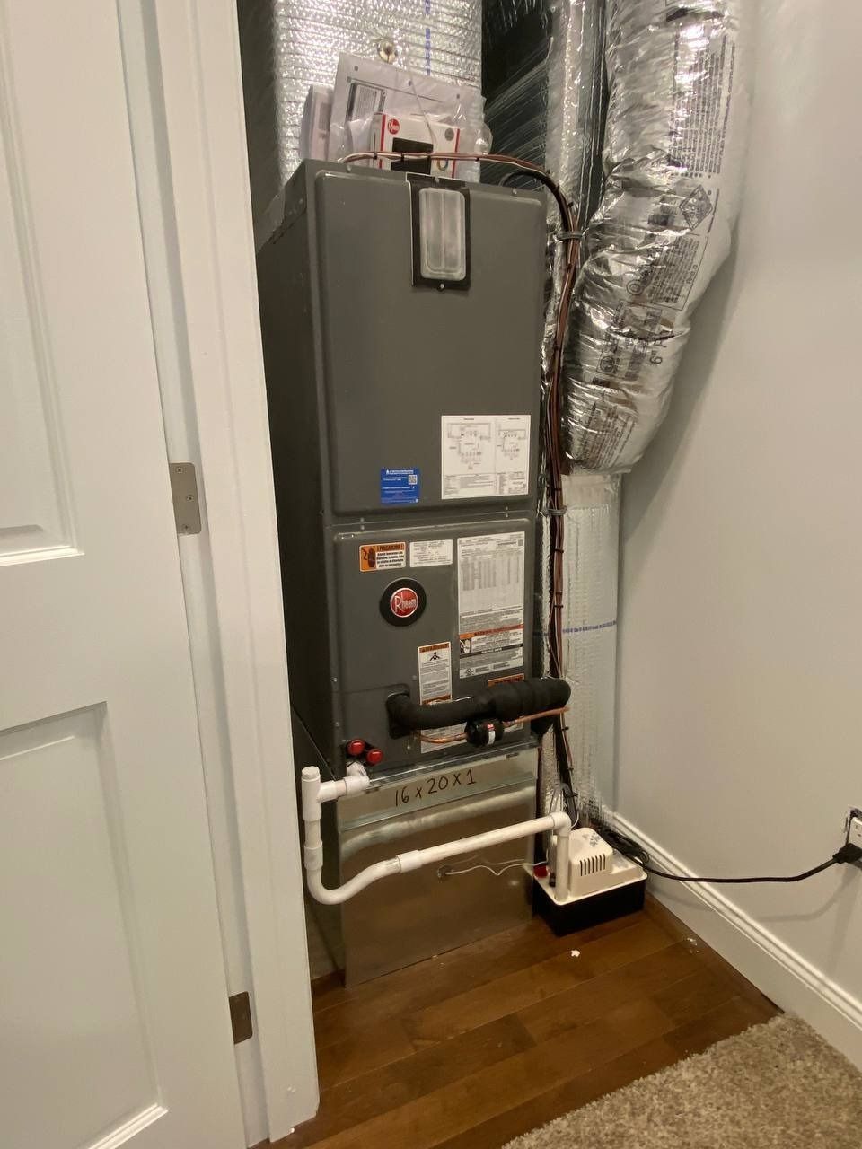 A furnace inside a white-walled closet, with ductwork and a white pipe system on a wooden floor.