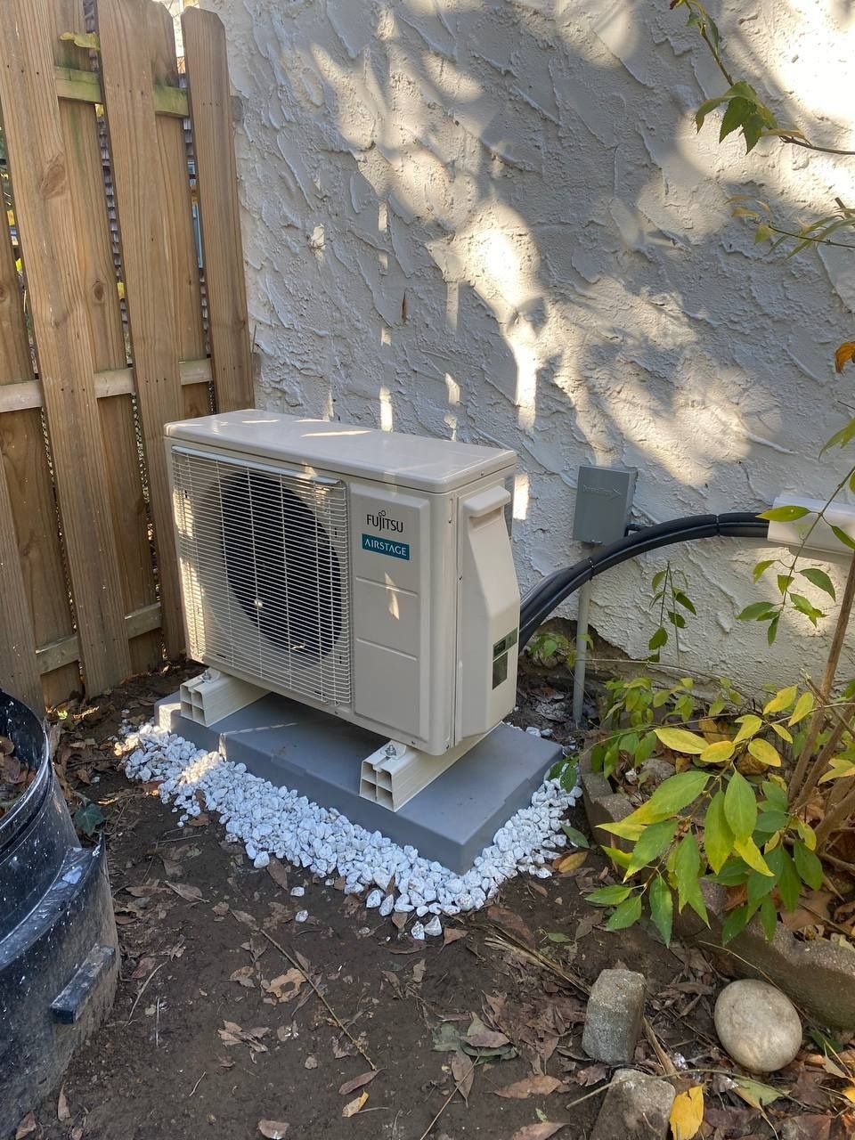 Air conditioning unit on concrete blocks, next to a fence and stucco wall. White rocks surround the base.