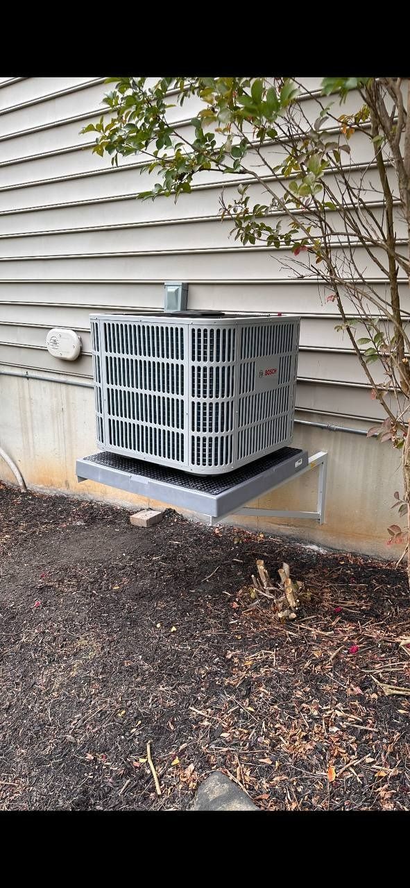 An air conditioning unit mounted on a metal platform attached to a house with dark mulch around it.