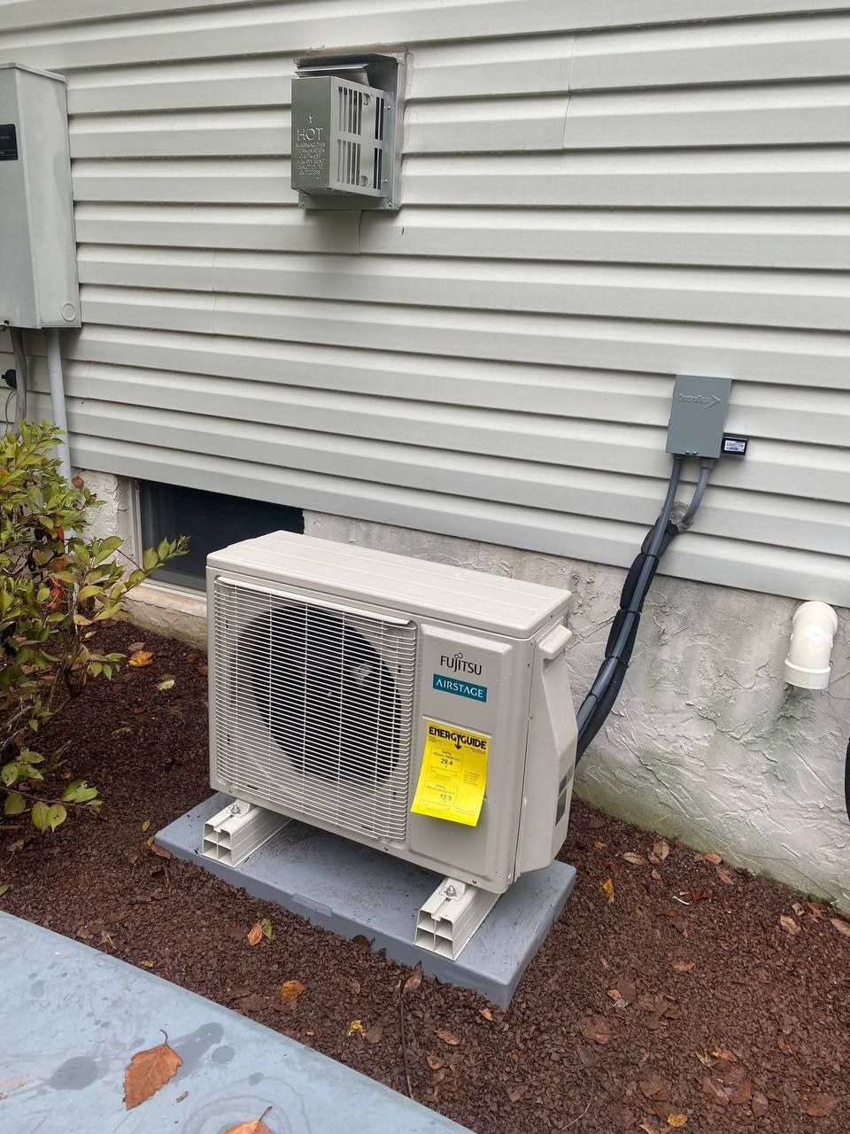 Outdoor heat pump unit mounted on a concrete pad next to a house with gray siding.