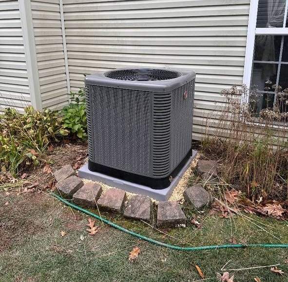 Central air conditioning unit outside a house, set on a concrete pad with surrounding brick border.
