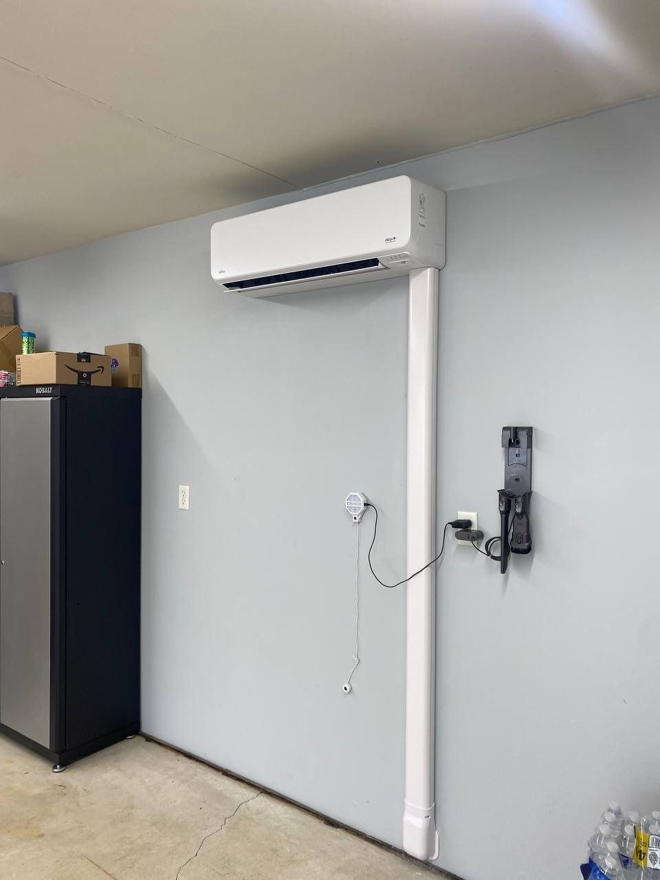 White air conditioning unit mounted on a gray garage wall. Power cords and outlet visible.