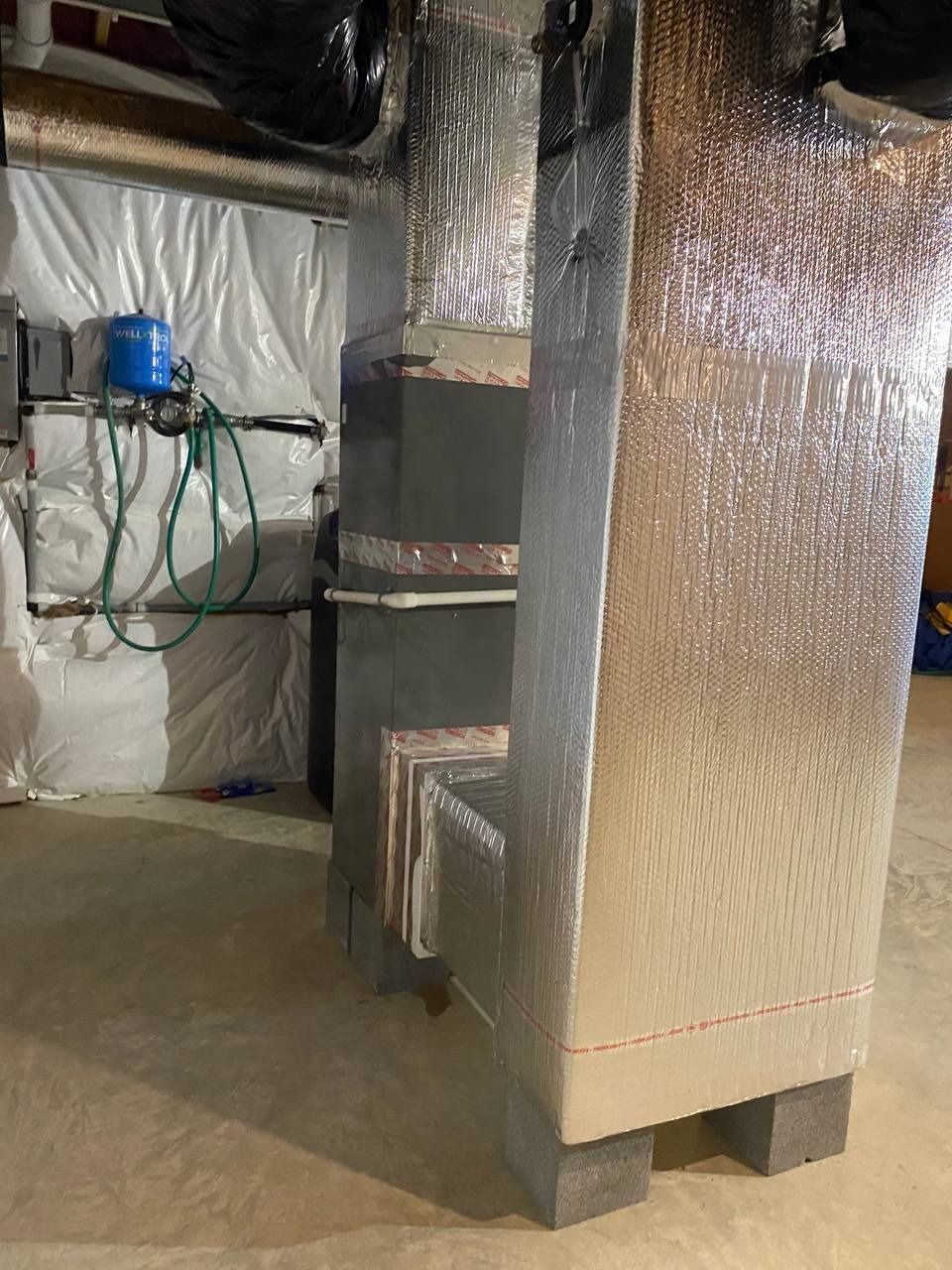 HVAC system covered in silver insulation in a basement setting. A blue water tank is visible in the background.