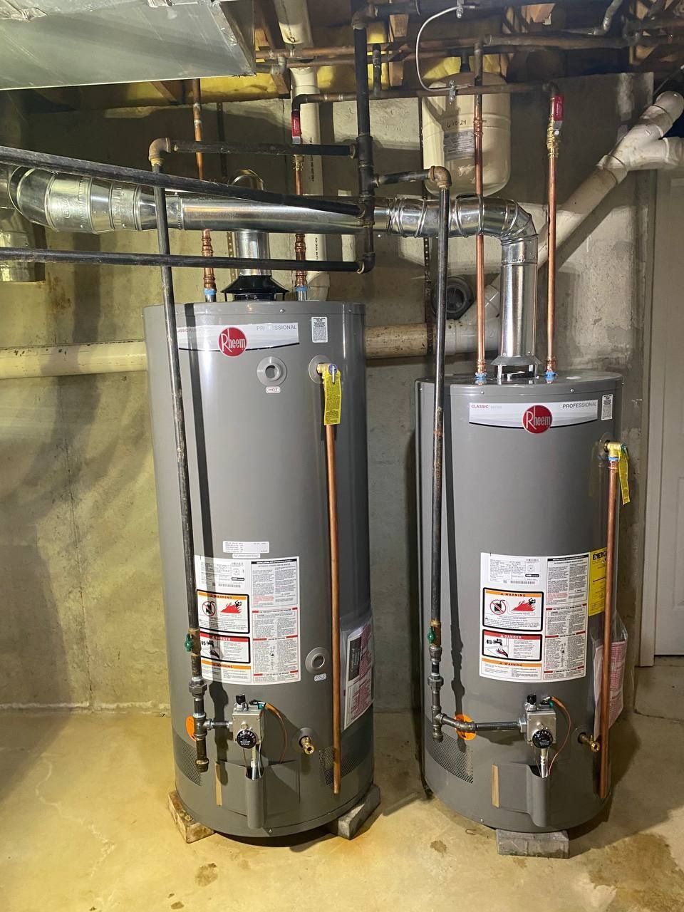 Two silver water heaters in a utility room, with copper pipes and a concrete wall.
