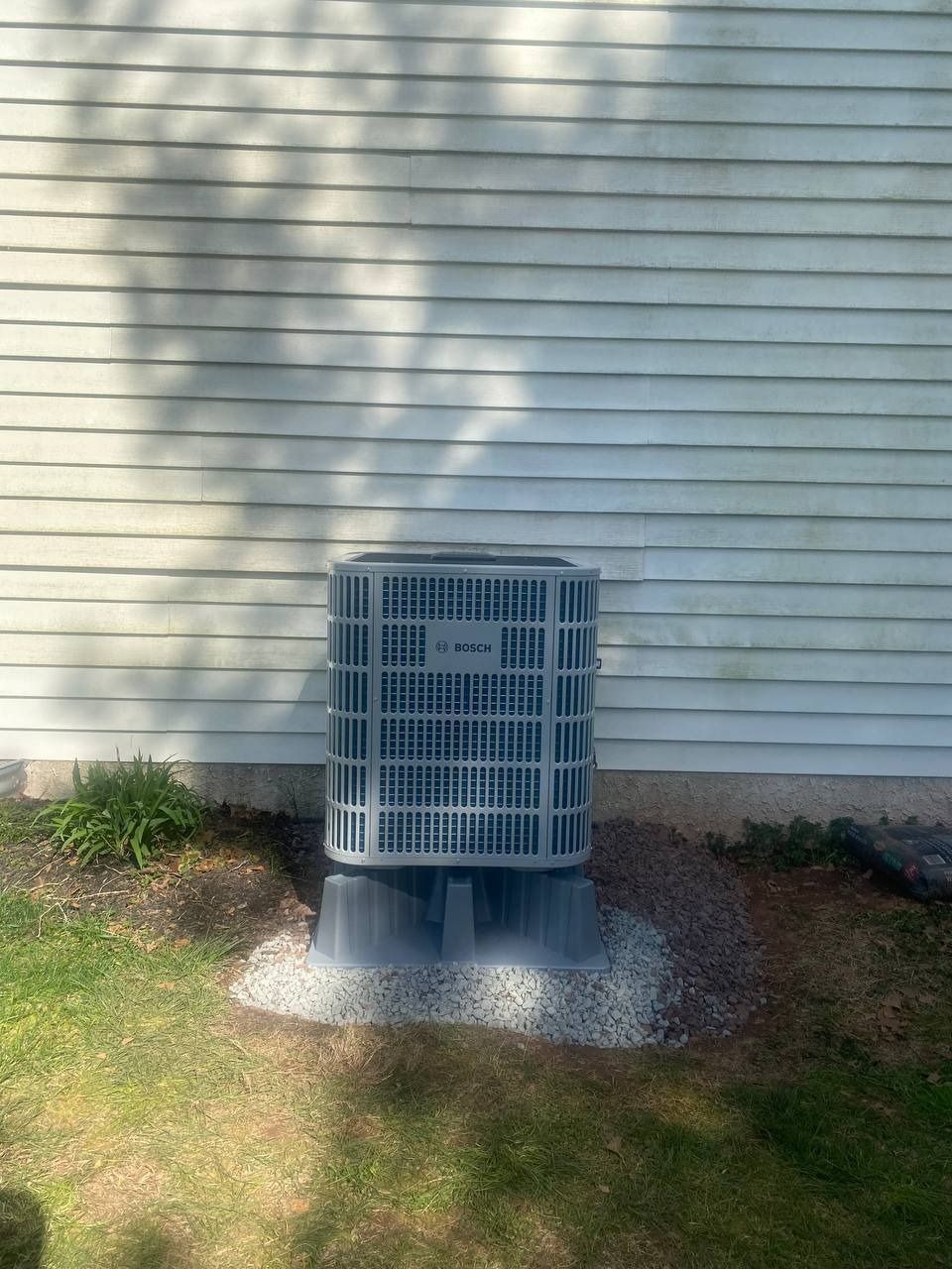 An outdoor air conditioning unit sits on a gravel base next to a white clapboard house.