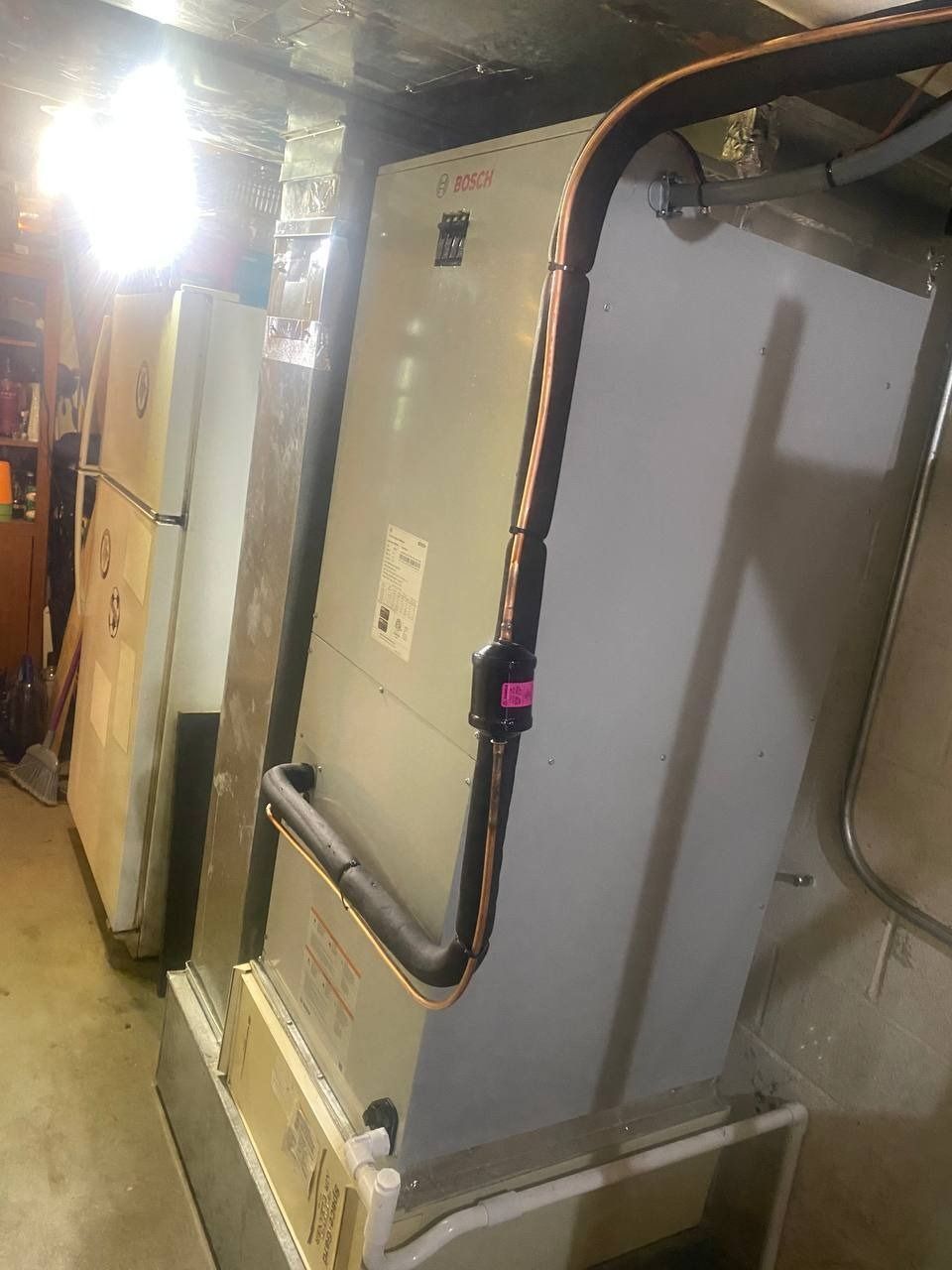 HVAC unit in a basement; silver with copper tubing, next to a white appliance, illuminated.