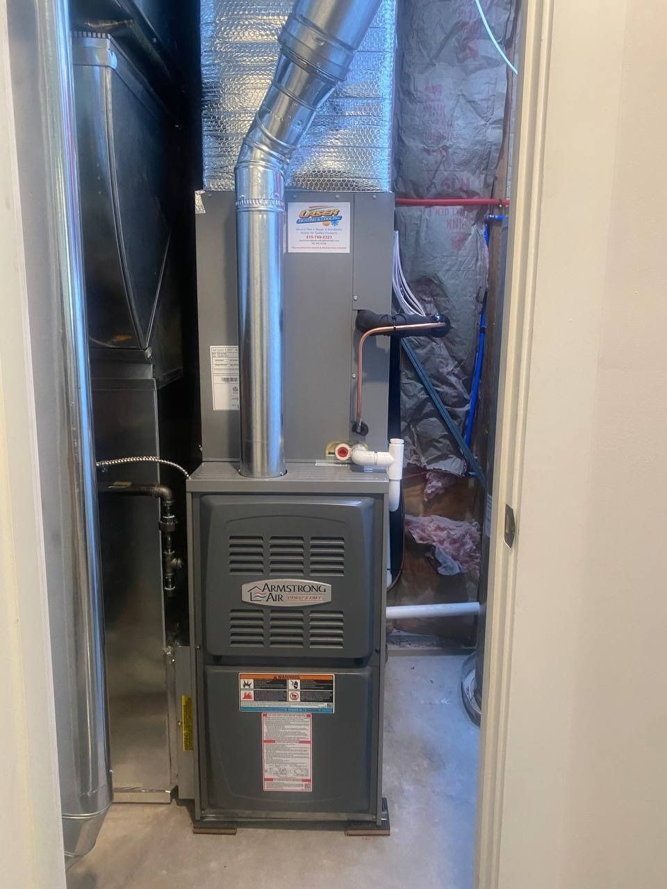 Furnace in a utility closet with ductwork and piping. Grey unit against insulated wall.