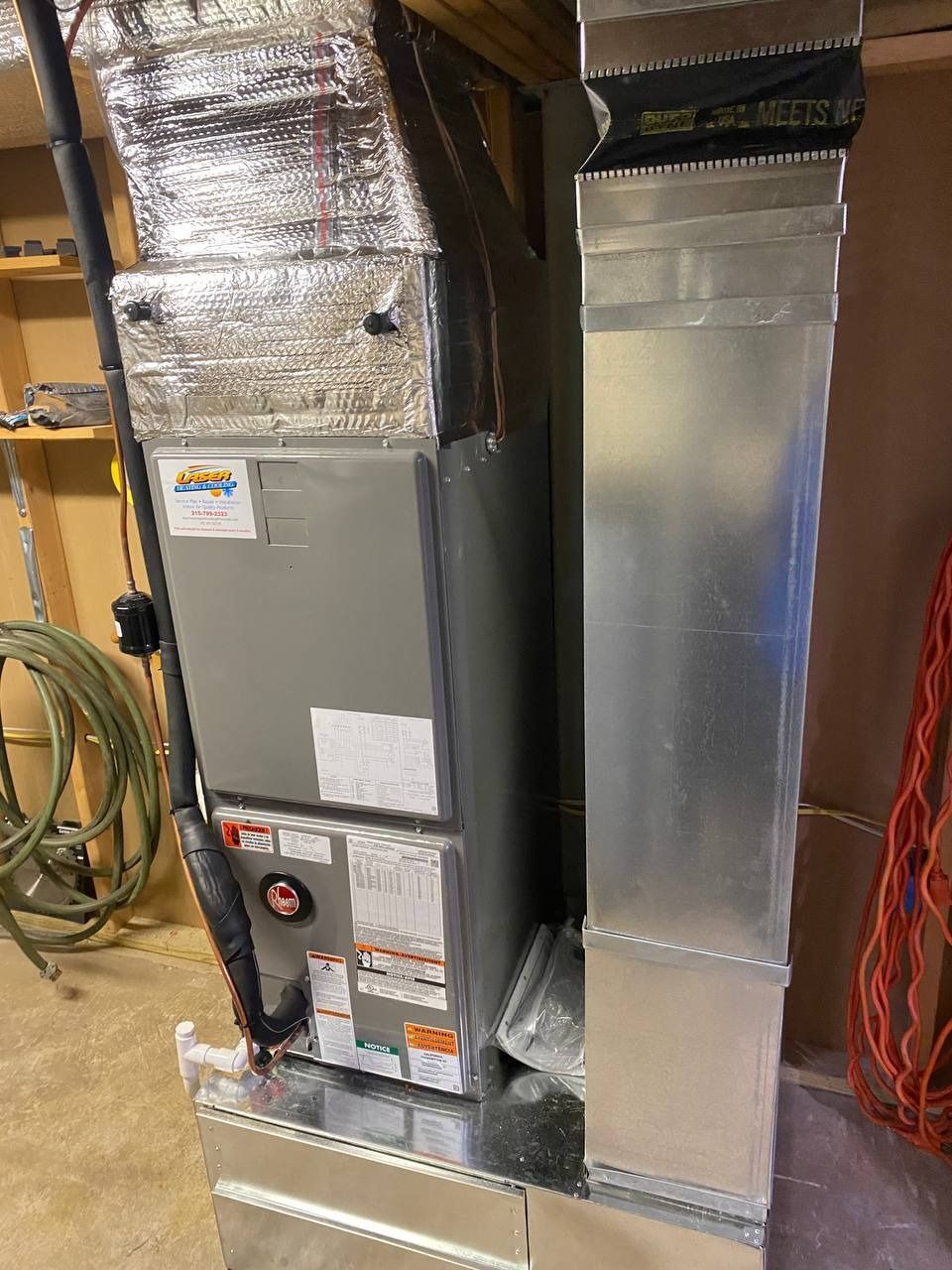 Furnace and ductwork in a utility room. Metal components, silver insulation, and various labels visible.
