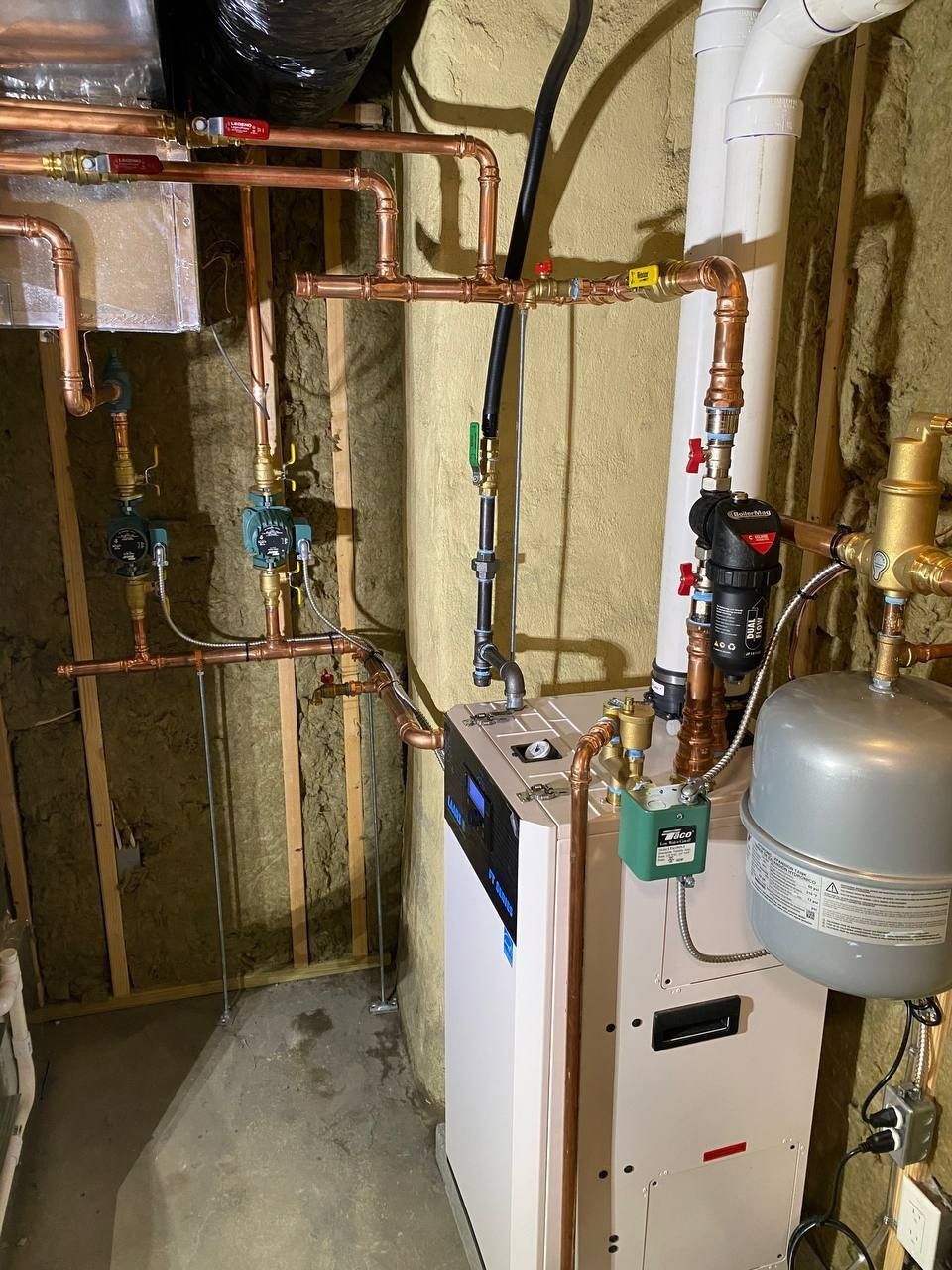 Copper pipes and valves connected to a white heating unit in a basement setting.