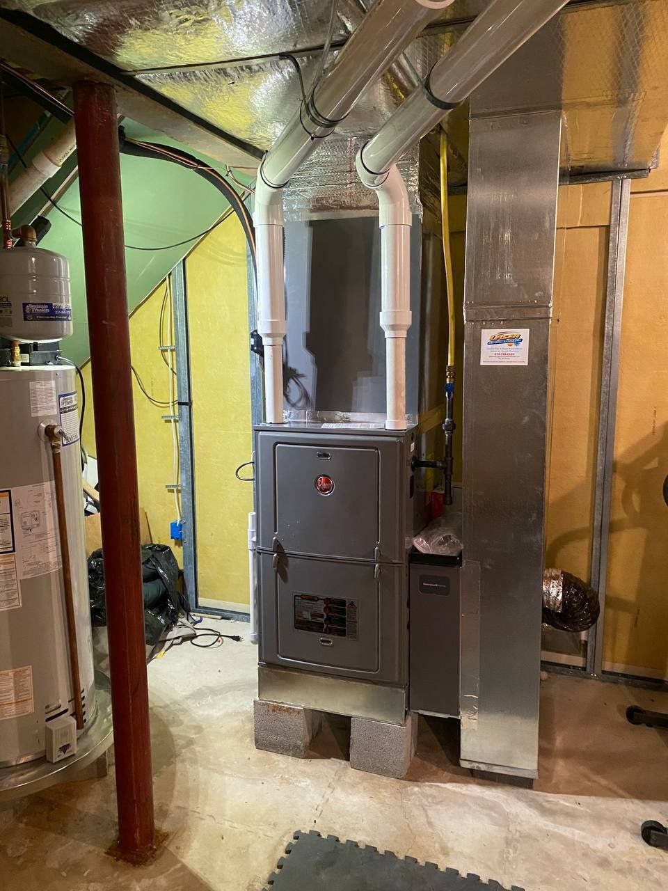 Furnace and water heater in a basement. The furnace is gray, with white vent pipes. A red support post is visible.