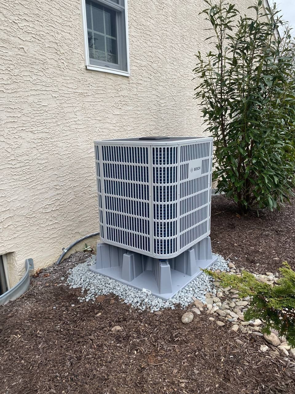 AC unit outside a building, on a gravel base, surrounded by mulch and bushes.