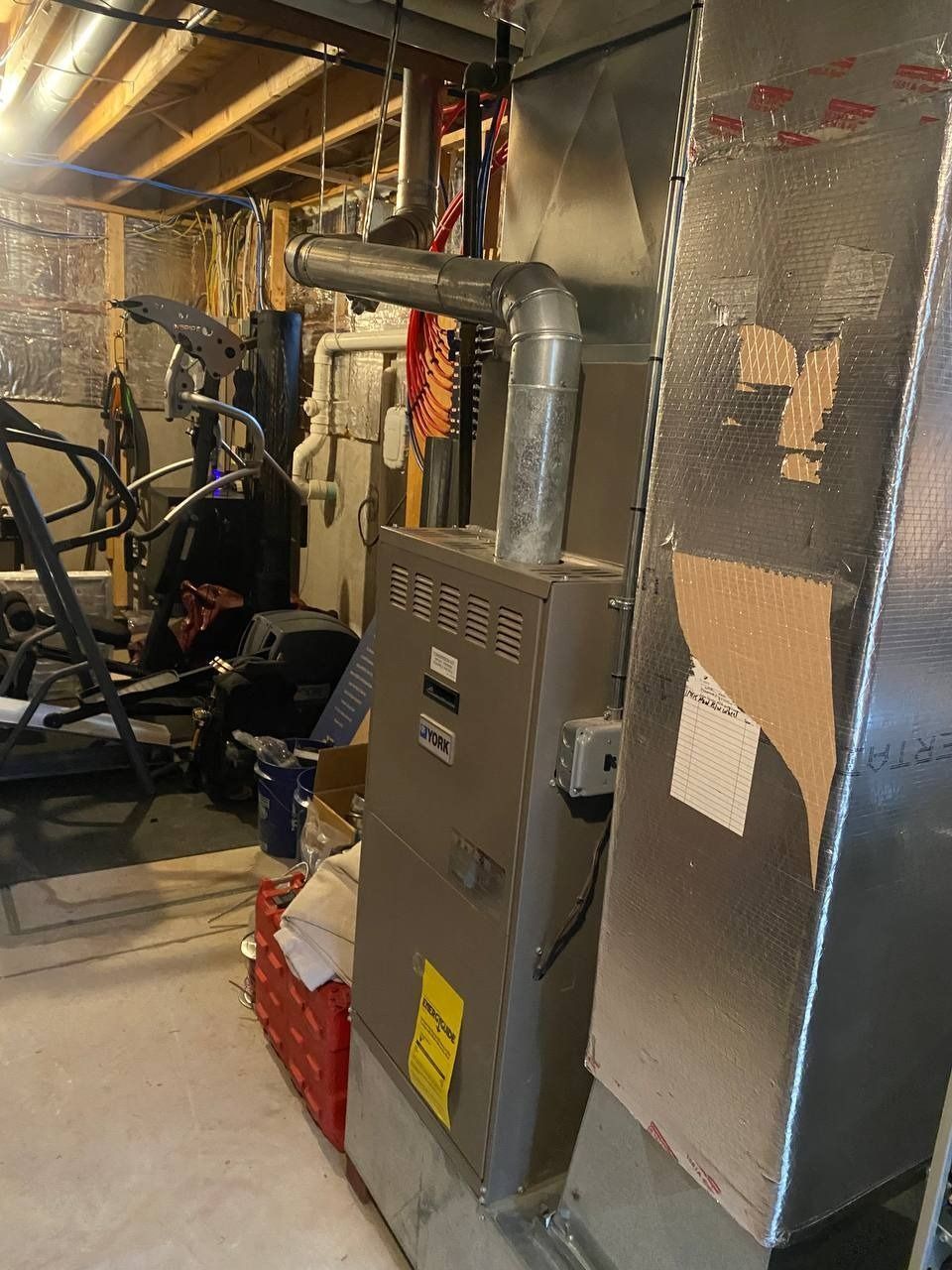 Furnace in a basement; silver metal with vent pipe. Yellow energy sticker on front.  Ductwork to the right.