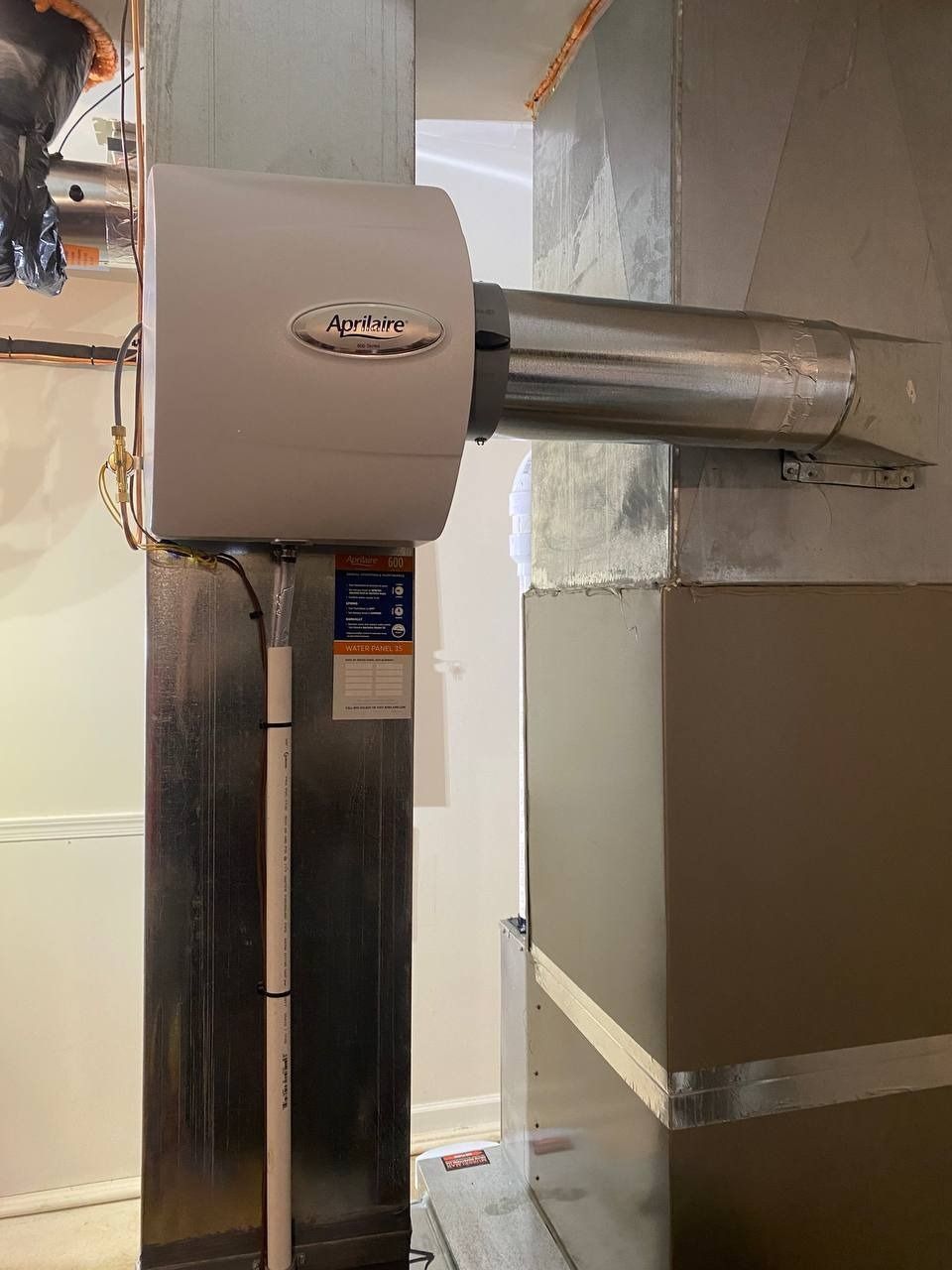 Humidifier attached to HVAC ductwork in a basement. White humidifier unit with silver ducting.