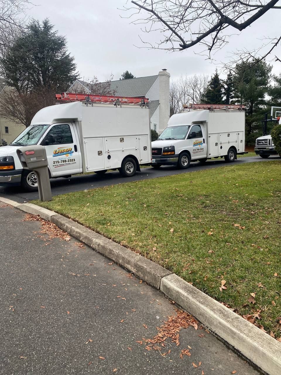 White service trucks parked on a residential driveway. They have ladders on top, grass and a mailbox are visible.