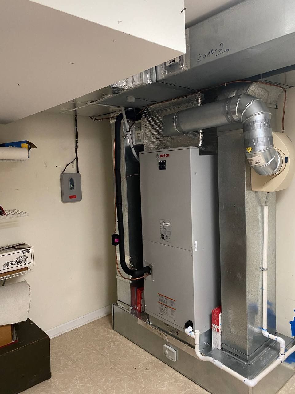 HVAC system in a basement. Silver furnace, ducts, and pipes against a beige wall. Gray electrical box on the left.