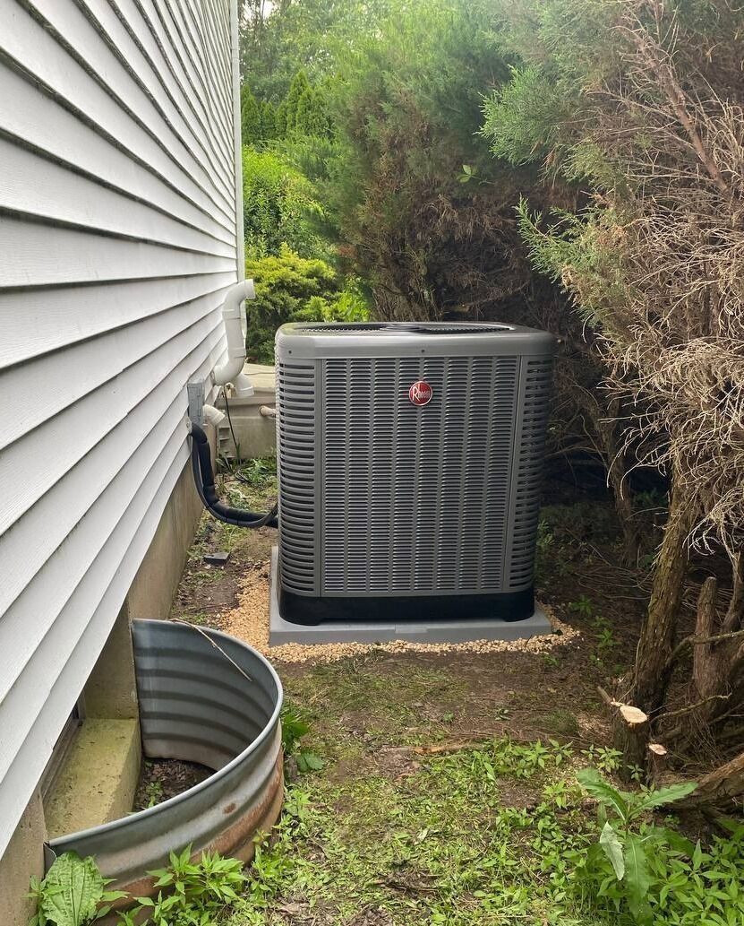 An air conditioning unit next to a house with a window well and overgrown foliage.