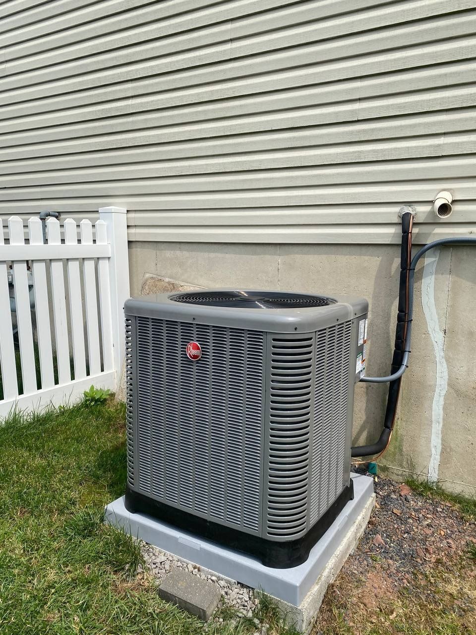 Air conditioning unit against a light gray building and white fence on a concrete pad.