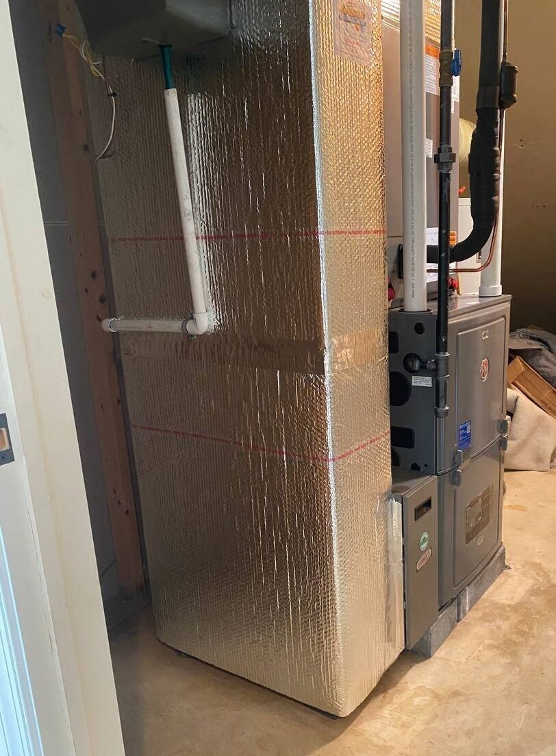 Furnace wrapped in silver insulation in a basement setting. White PVC pipes and black gas line visible.