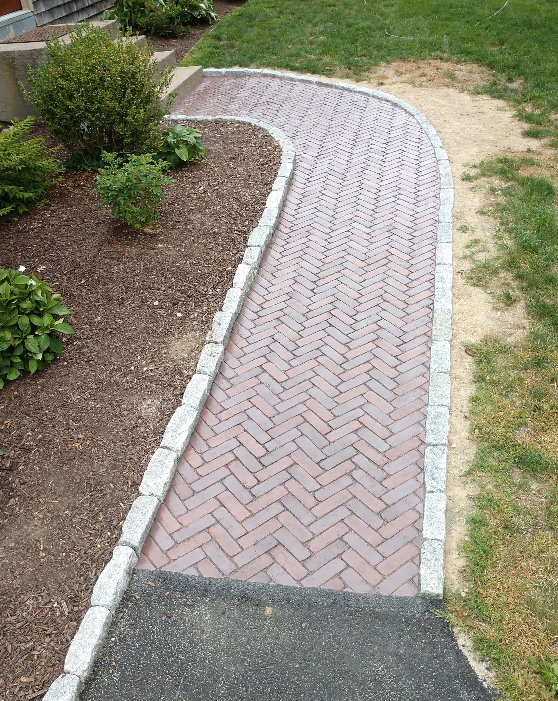 A brick walkway with a white border is surrounded by grass and bushes.