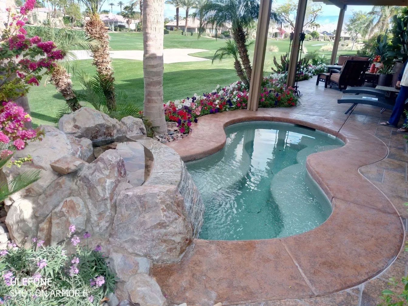 A small swimming pool surrounded by rocks and flowers in a backyard.