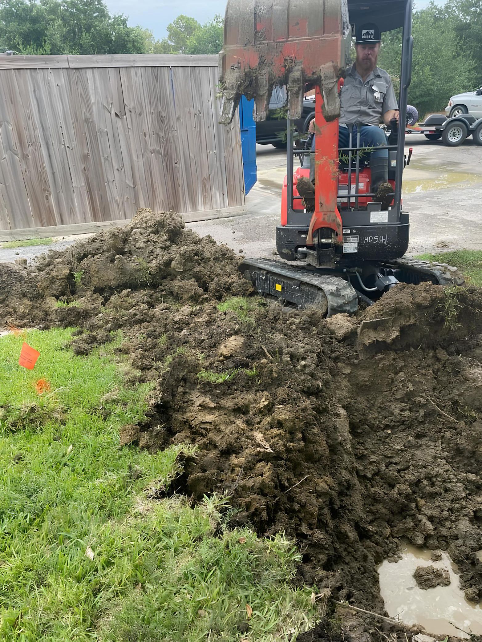 A person operating an excavator digs a trench in a grassy area, with a pile of dirt nearby.