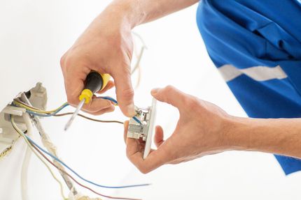 Electrician installing an electrical outlet with a screwdriver; white wall, blue uniform.