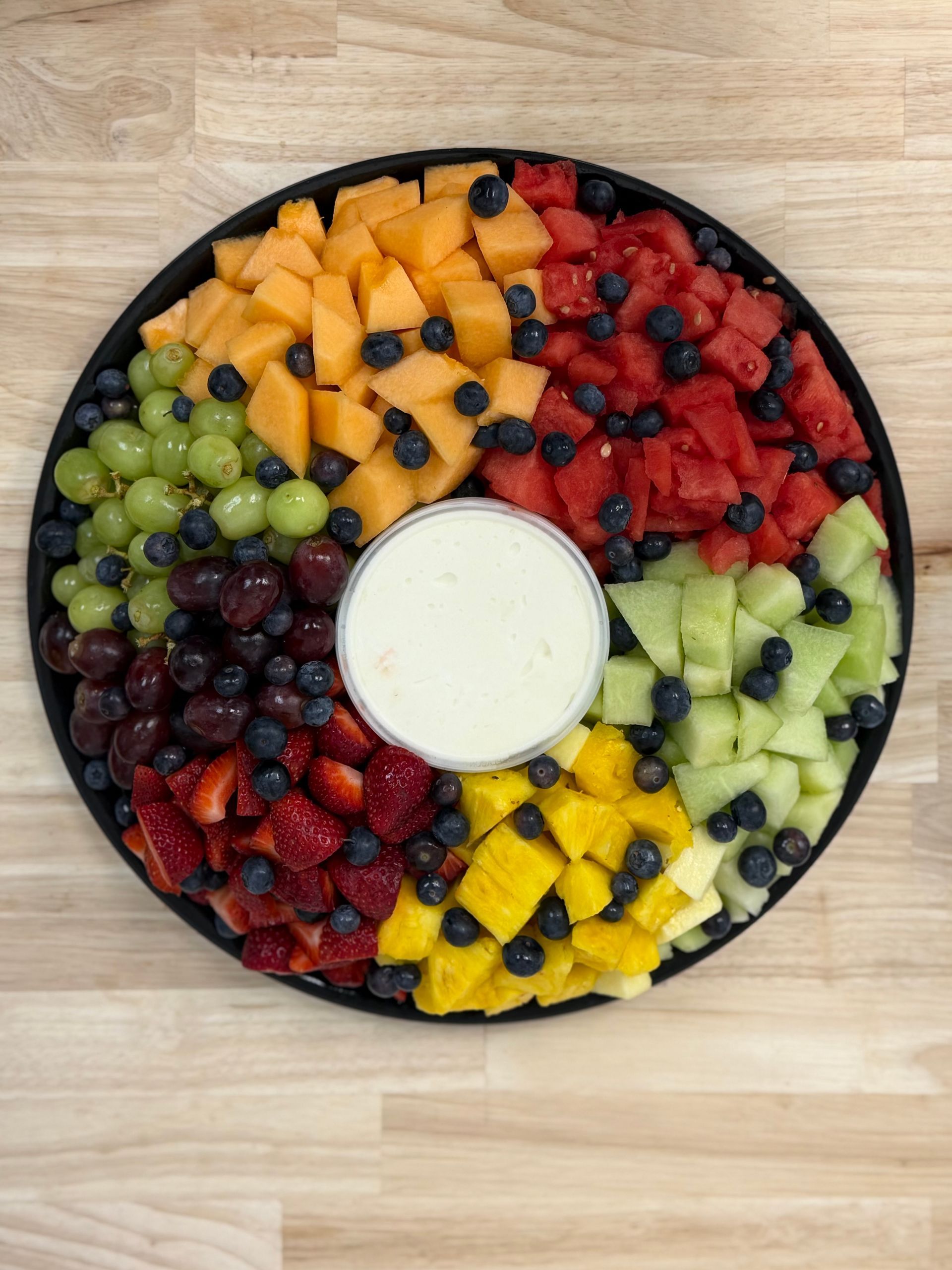 Fruit platter with dip in the center, arranged in colorful sections on a round black tray.