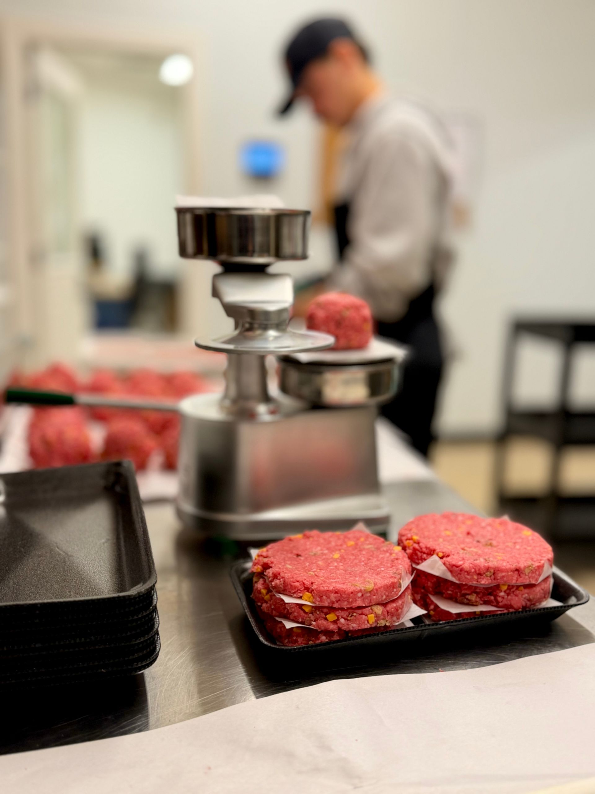 Raw burger patties on a counter beside a meat grinder, with a blurred cook in the background