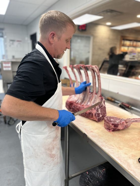 Butcher trimming a large cut of raw meat on a counter, wearing a white apron and blue gloves.
