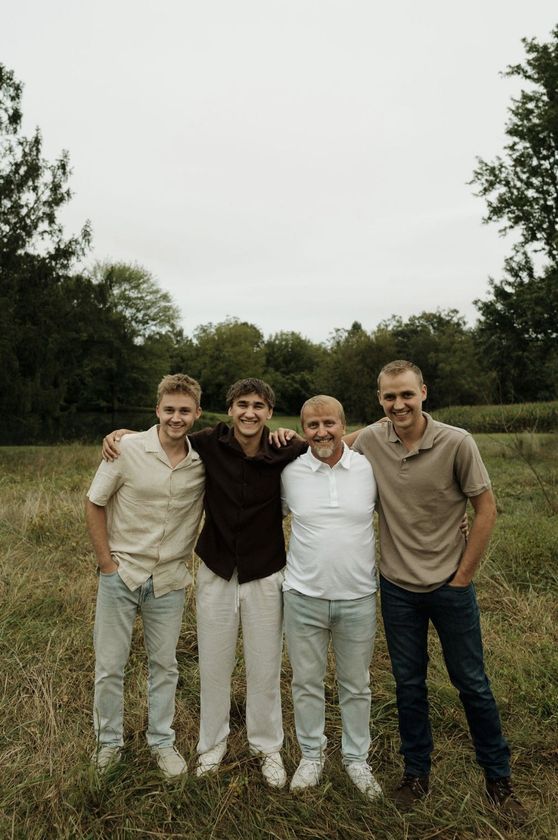Four people standing in a field with arms around each other, smiling. Cloudy sky, trees in the background.