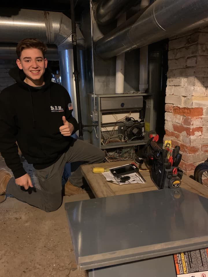 A young man is kneeling down in front of a furnace and giving a thumbs up.