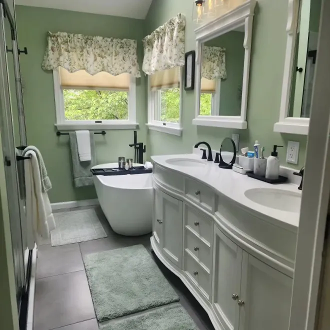 A bathroom with a white tub, double sink, and green walls; sunlight streams in from windows.